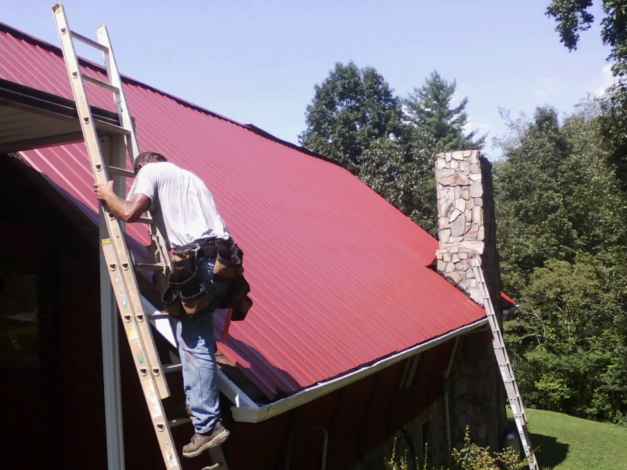 Roofer on a ladder beside a red metal roof and stone chimney — Soffit & Fascia Repair by Roofing Brothers in Hamden